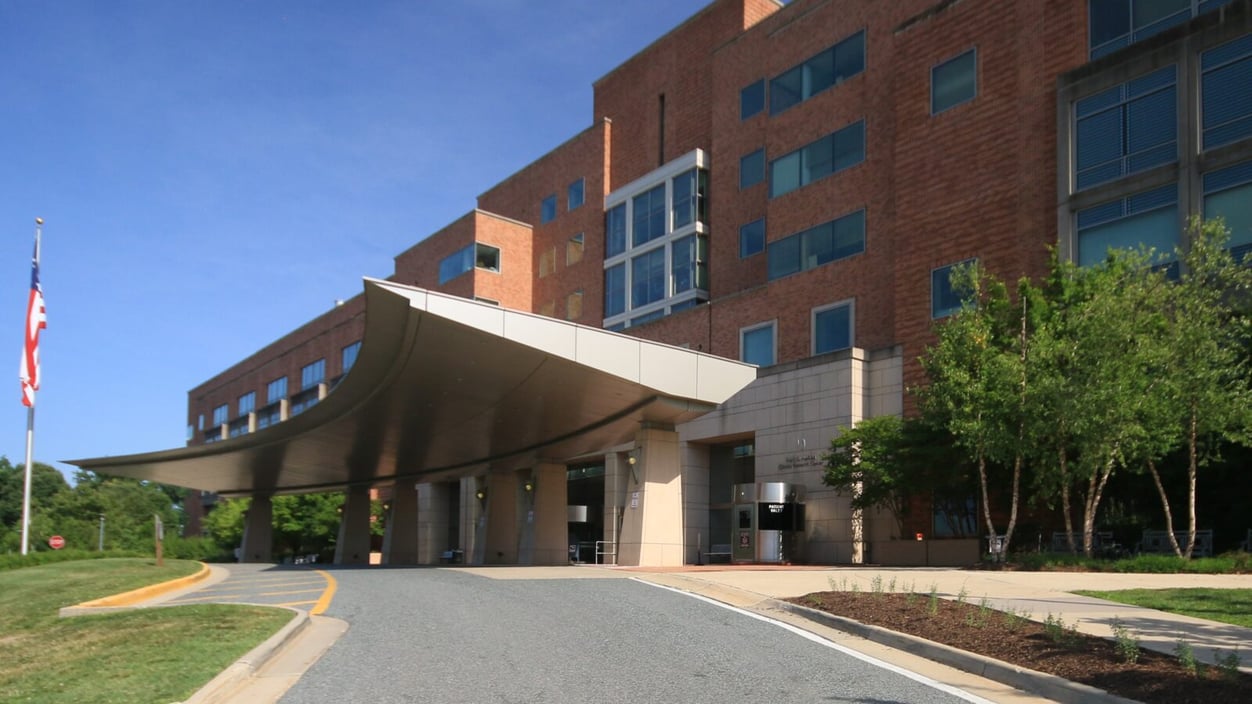 The front of the NIH Clinical Center in Bethesda, MD. It's a brick building with a large, curved metal overhang over the front doors.