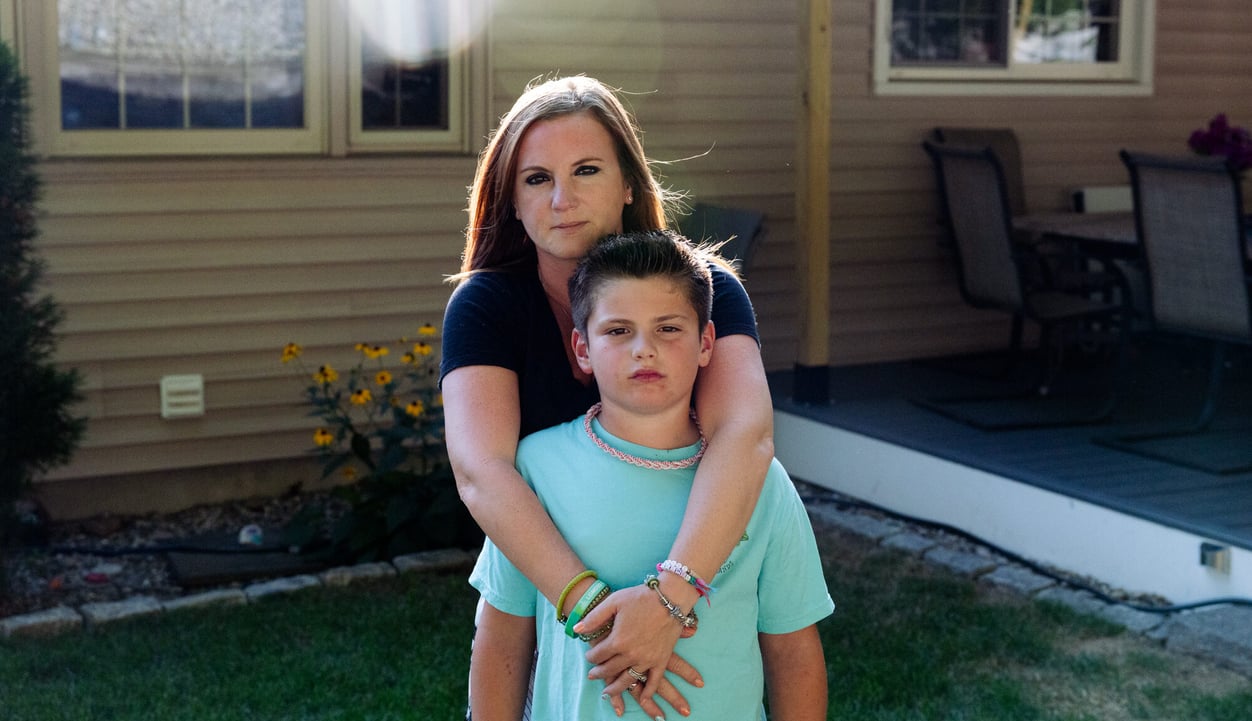 A mother hugs her son from behind, both looking straight into the camera