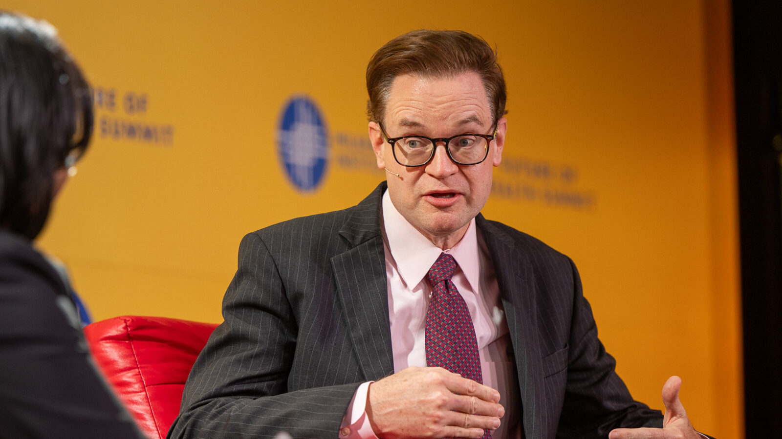 Jim O'Neill speaks at a conference sitting in a suit on a red armchair
