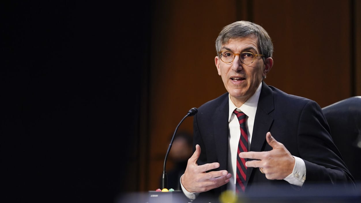 Peter Marks sits at a microphone. He's a white man with grey hair, wearing orange tinted glasses and a suit with a red and blue striped tie.