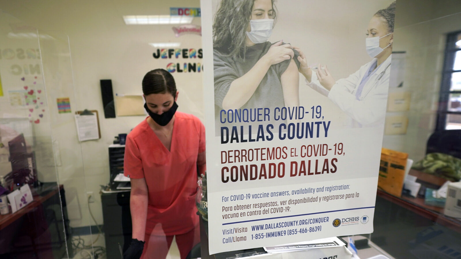 A nurse administers a Covid shot, as viewed through a glass wall, behind a poster advertising the shots. 