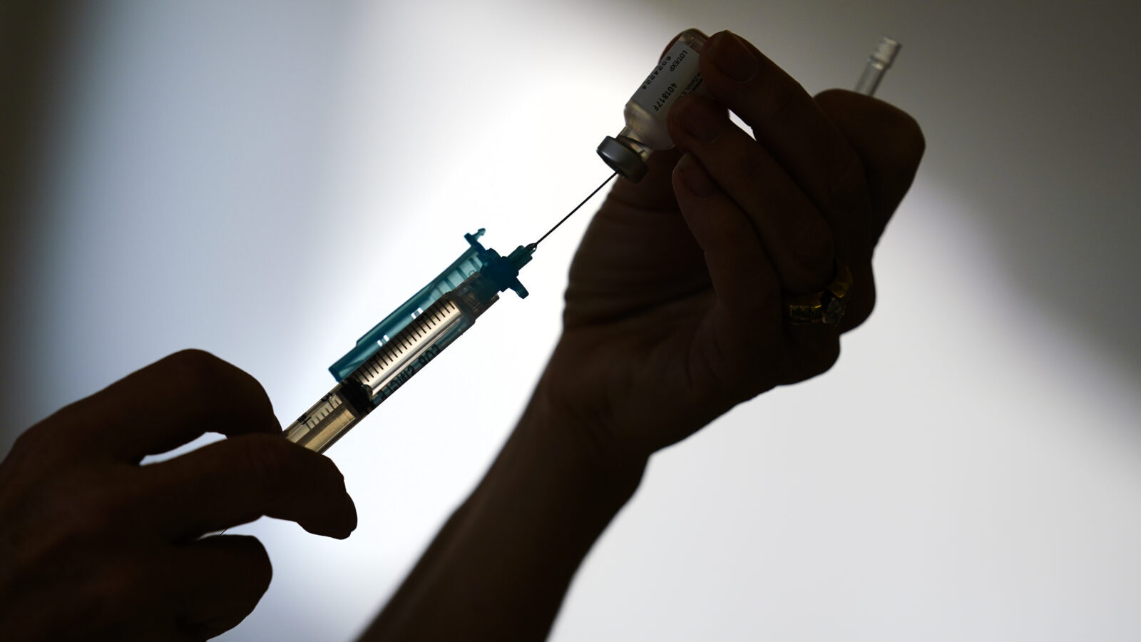 A hand in shadow holds a vaccine syringe up, with the vial attached and facing down.
