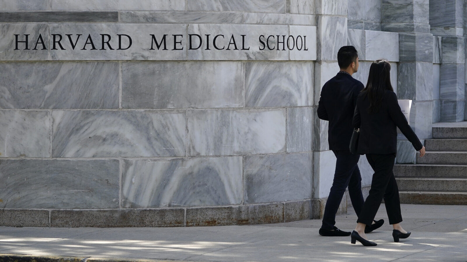 Two people walk away from view, next to a large marble wall with lettering on it that says "Harvard Medical School."