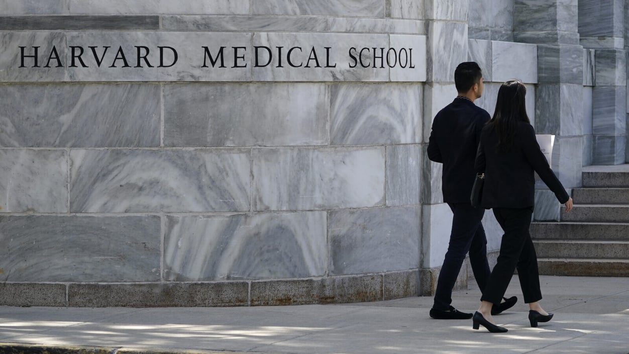 Two people walk away from view, next to a large marble wall with lettering on it that says "Harvard Medical School."