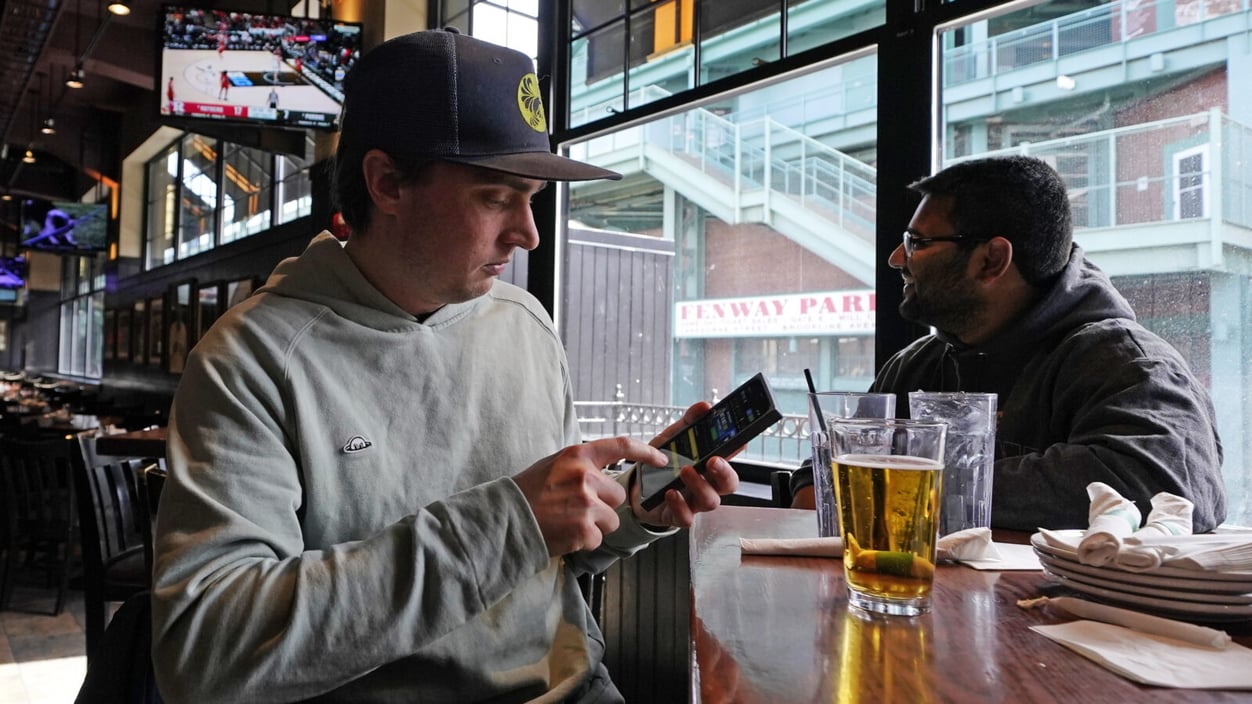 Taylor Foehl, a young man in a grey sweatshirt and flat-ish rim baseball hat, looks at the mobile betting app on his phone after placing a wager on a men's college basketball game at a Boston sports bar in 2023