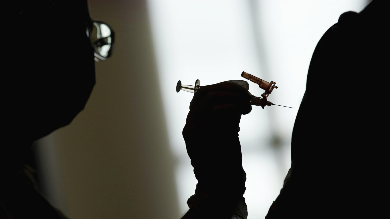 A dark figure poises a needle over what resembles a person's arm. They are both silhouetted by a window in the background.