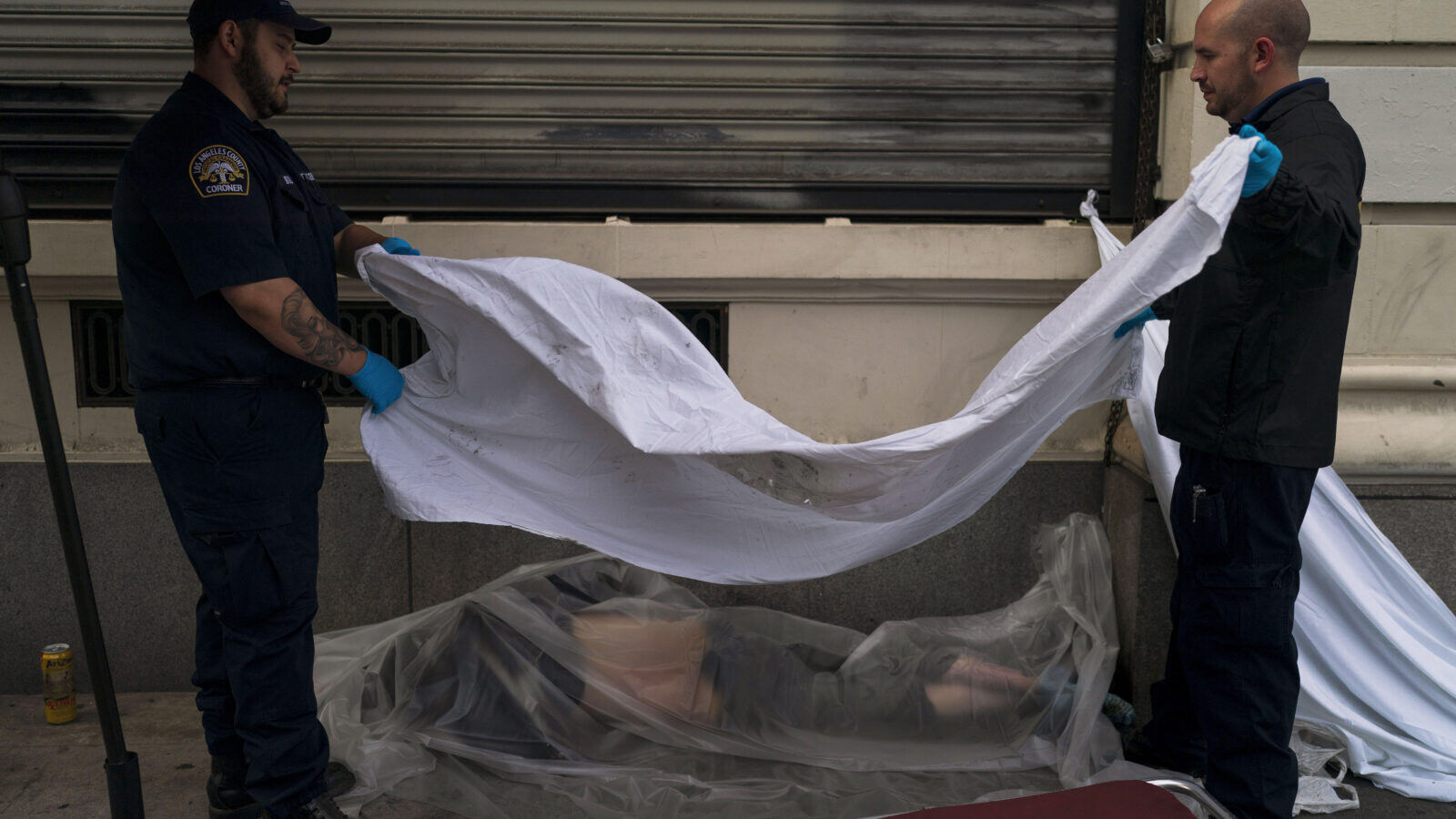 Two emergency responders drape plastic and a white cloth over a man who was found dead on the sidewalk after a drug overdose.