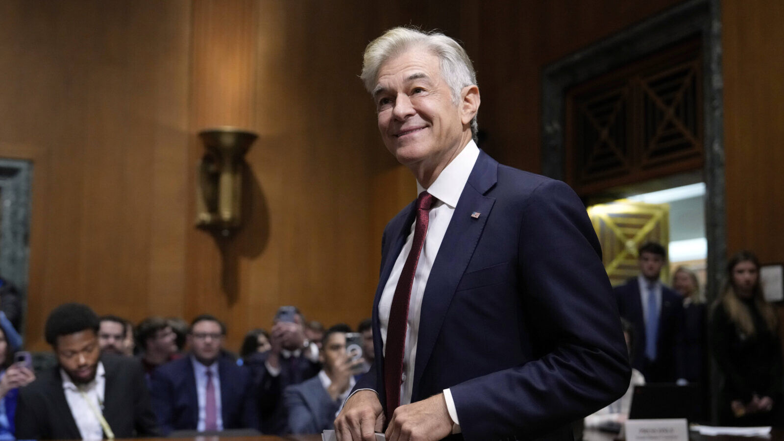 Dr. Oz, in a blue suit with light white hair, stands at his Senate committee hearing on Friday.