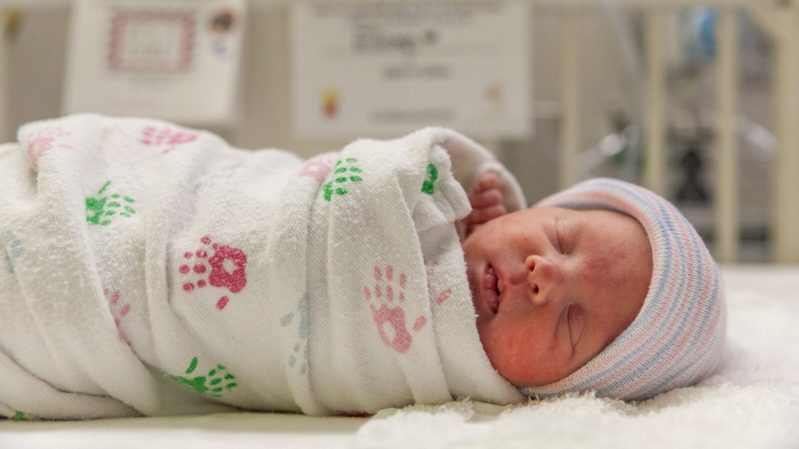 A picture of a sleeping infant wrapped in a white blanket with colored handprints and a pink-and-blue-striped hate