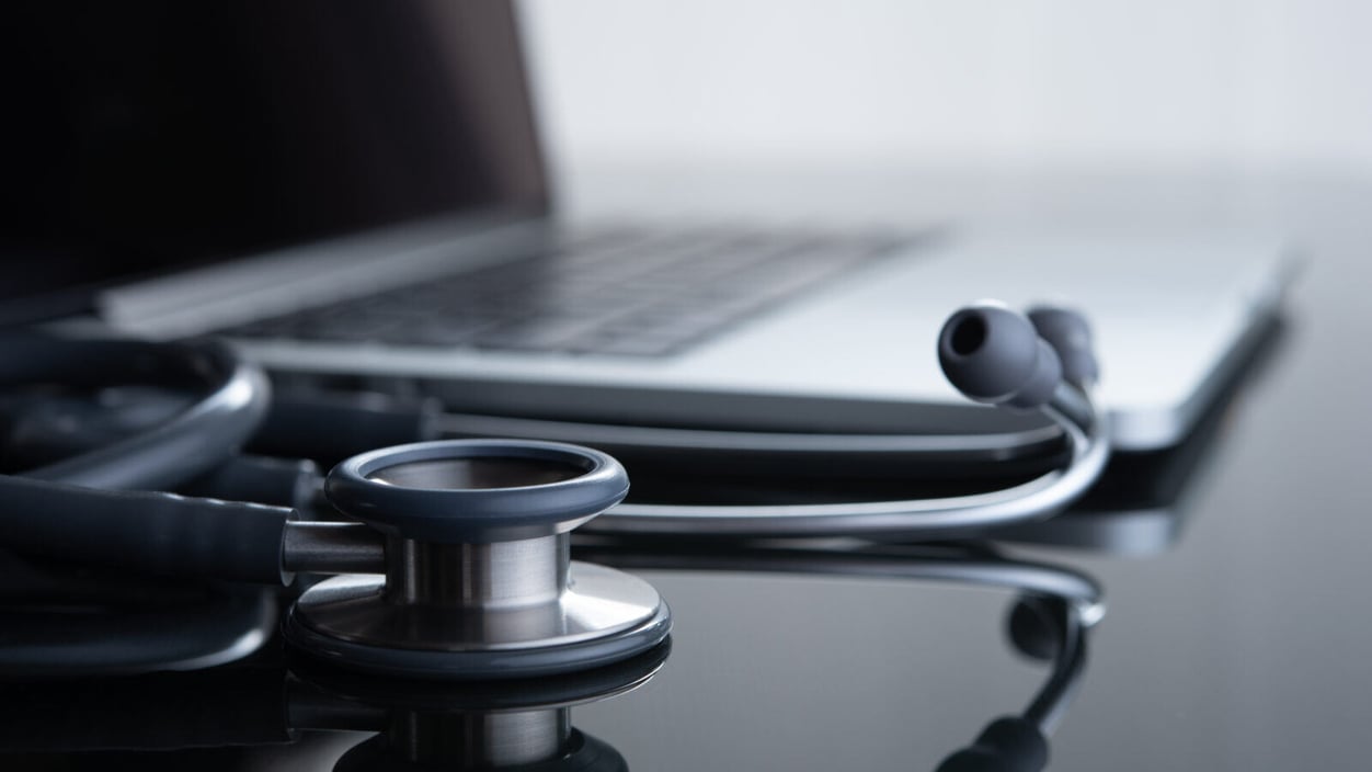 A stethoscope lays on a glass table next to a laptop.