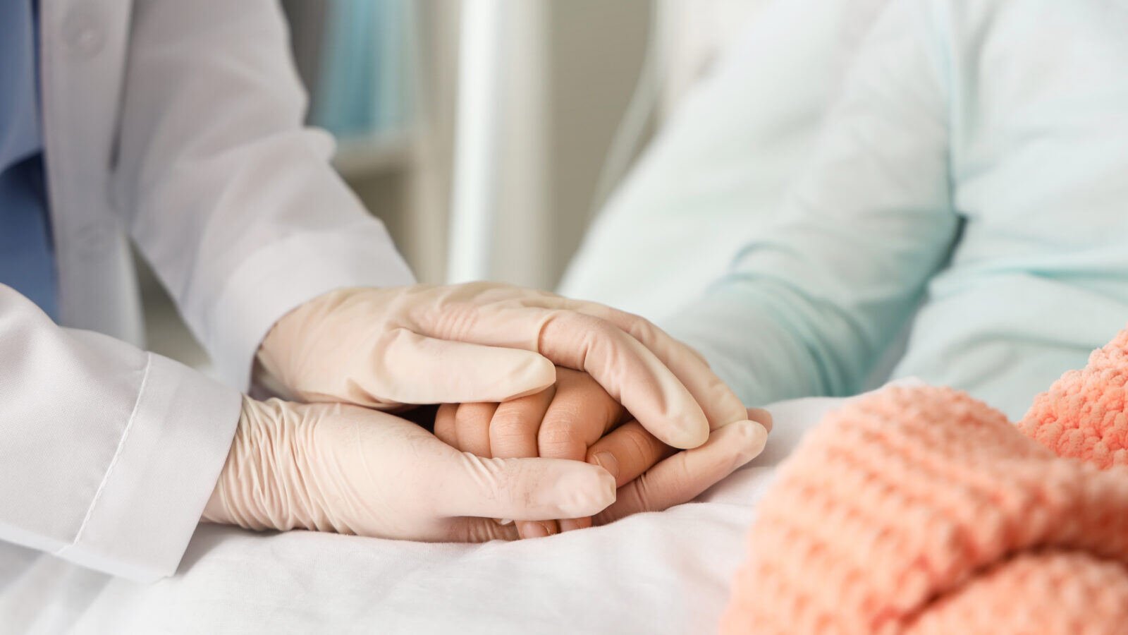 A close up image of a clinician's gloved hands holding a child's hand. The child is in a hospital bed. 