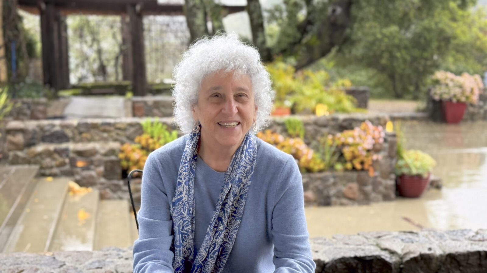 Marion Nestle,  an octogenarian with a cloud of curly white hair, sits in a garden smiling at the camera.