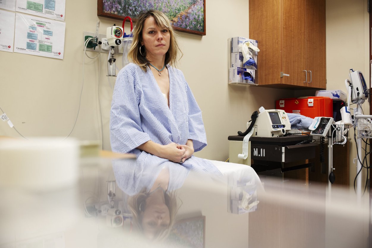 Anna Rathkopf sits in a hospital room with a blue striped medical gown wrapped around her. She stares solemnly into the camera.