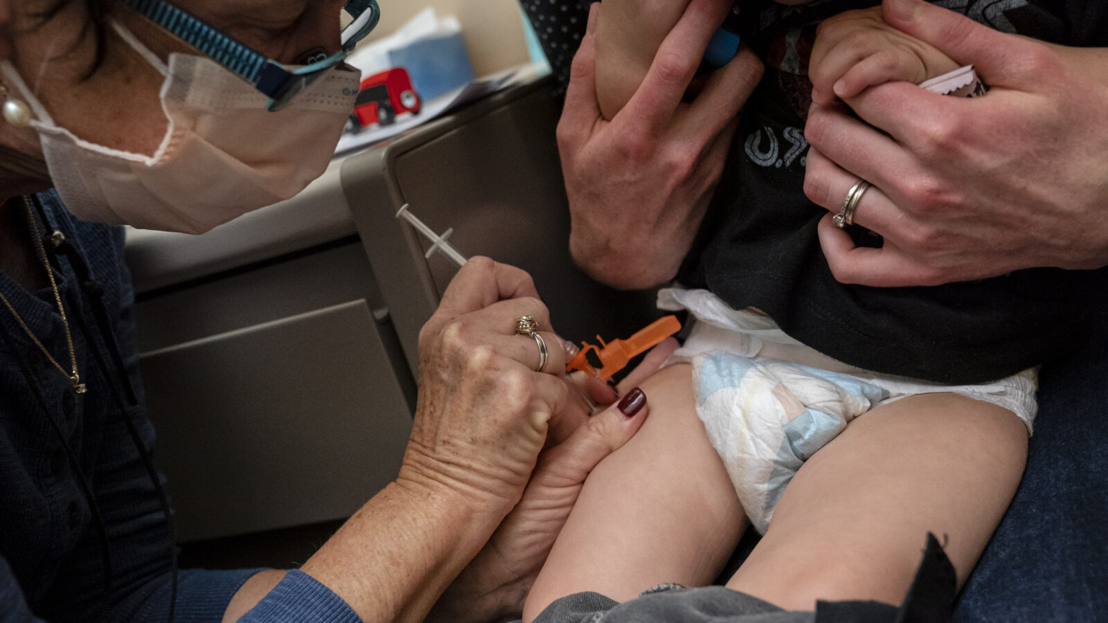 A toddler receives a vaccine in the leg from a masked clinician.