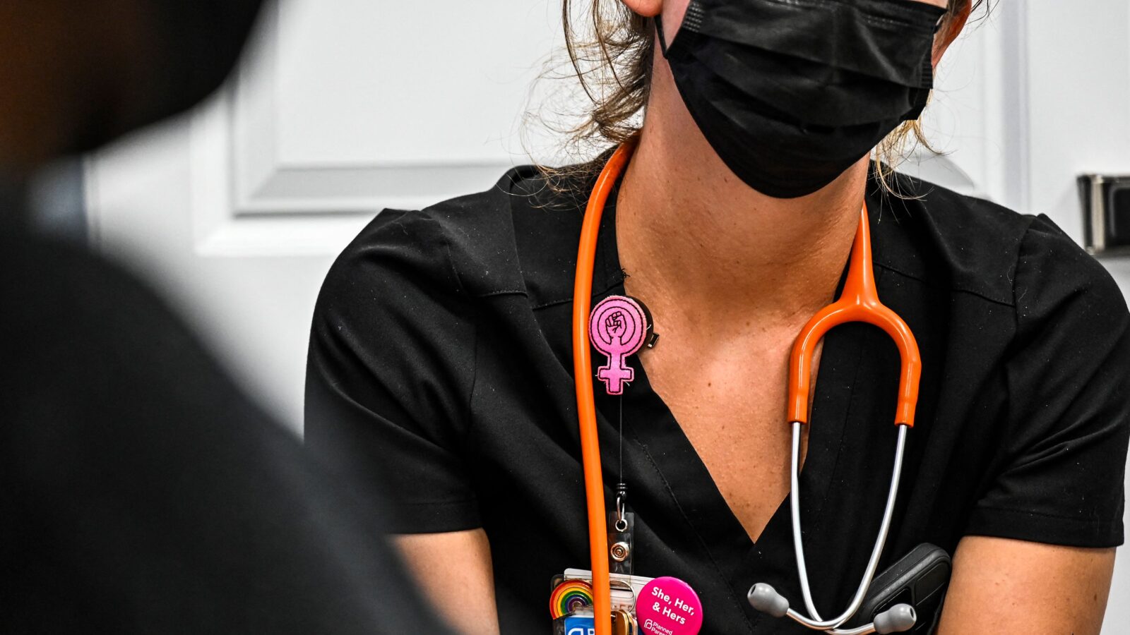We see the torso of a clinician wearing black scrubs, an orange stethescope, and a pink gender equity pin.