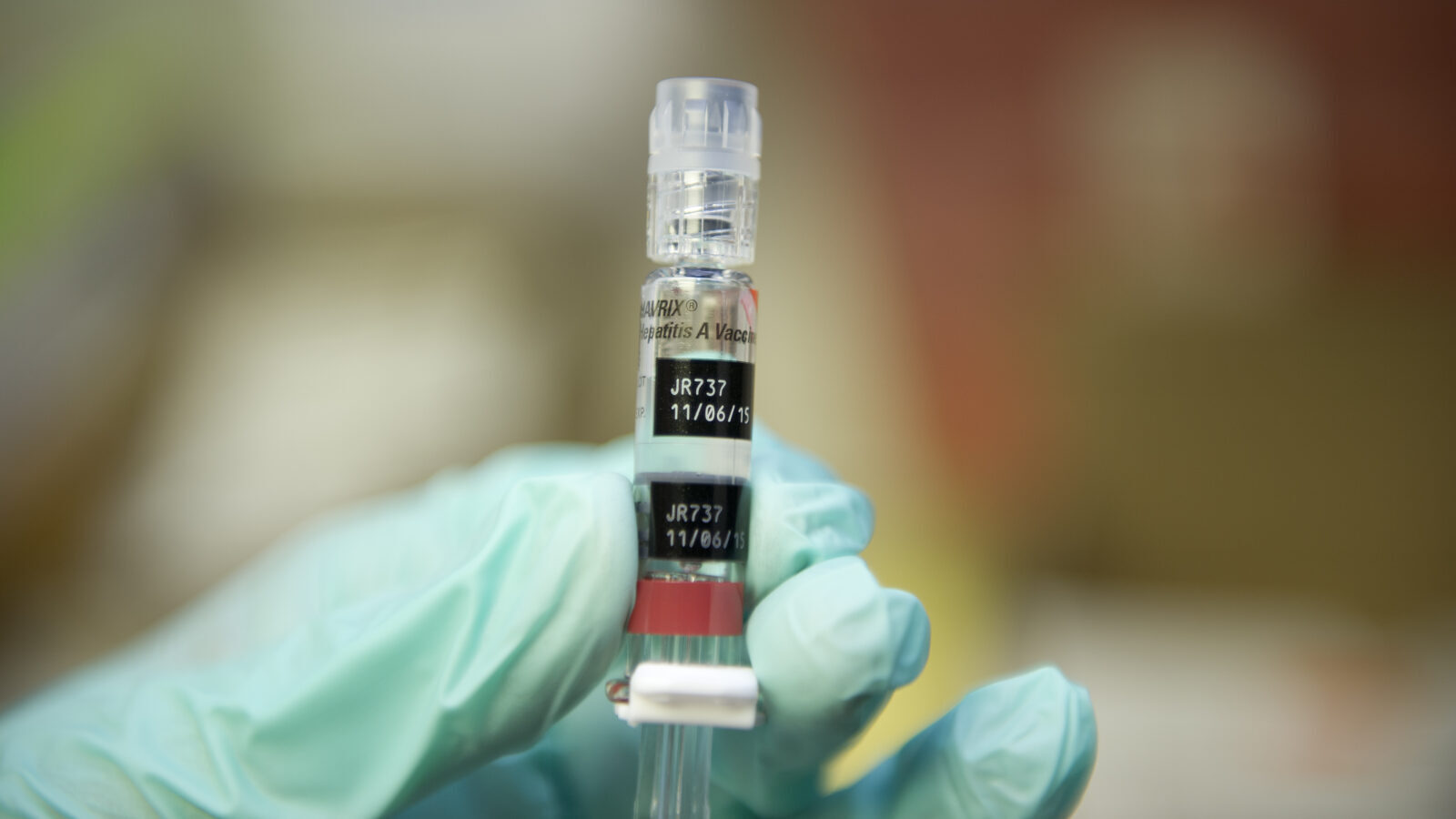 A gloved hand holds up a syringe with a hepatitis B vaccine