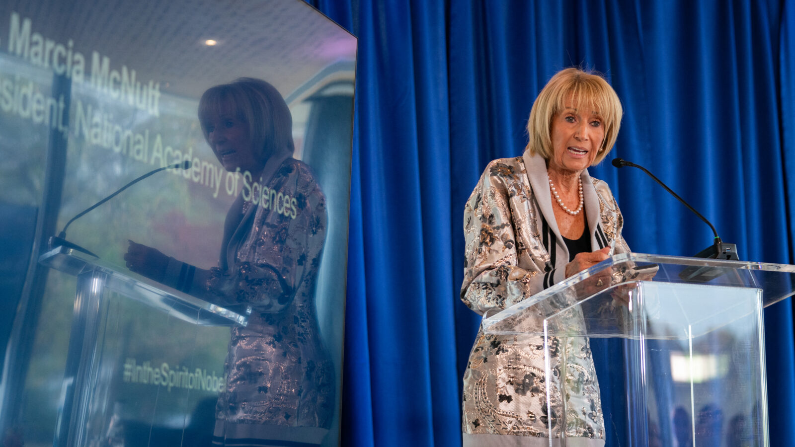 Marcia McNutt stands at a clear glass podium, speaking during the 2024 Nobel Prize Symposium at the Swedish Embassy in Washington.