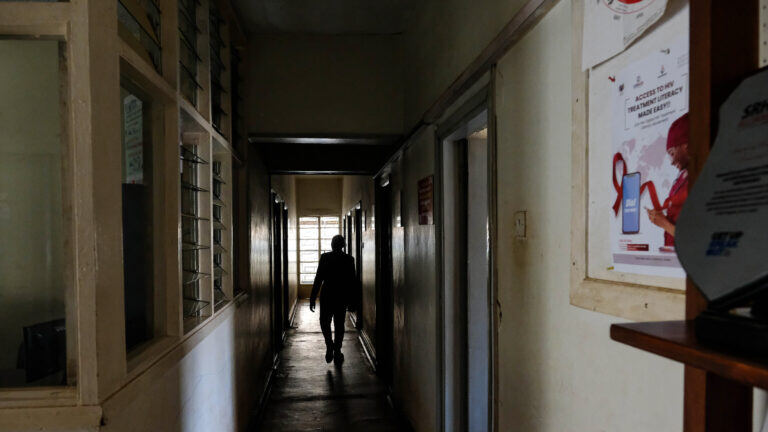 A person walks down an empty aisle illuminated by dim sunlight from behind. A flyer displaying the red HIV/AIDS awareness ribbon is on the right wall 