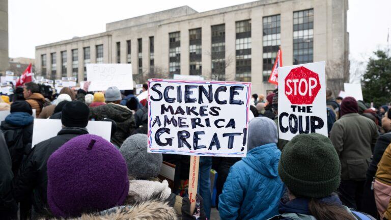A crowd rallies outside the Health and Human Services headquarters in Washington. One sign reads "Science makes America great," and another reads "Stop the Coup" but the word "stop" is painted like a stop sign.