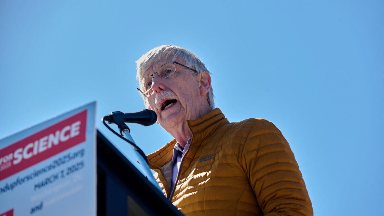 Francis Collins, an older man with all-white hair, speaks at a podium with a clear blue sky behind him