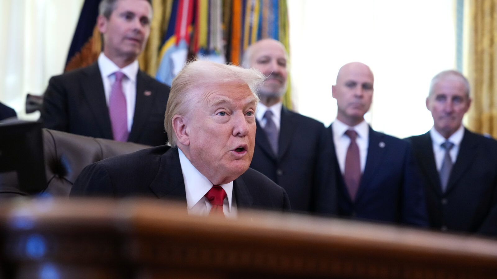 Trump at his desk, with four out-of-focus men in suits standing behind him.
