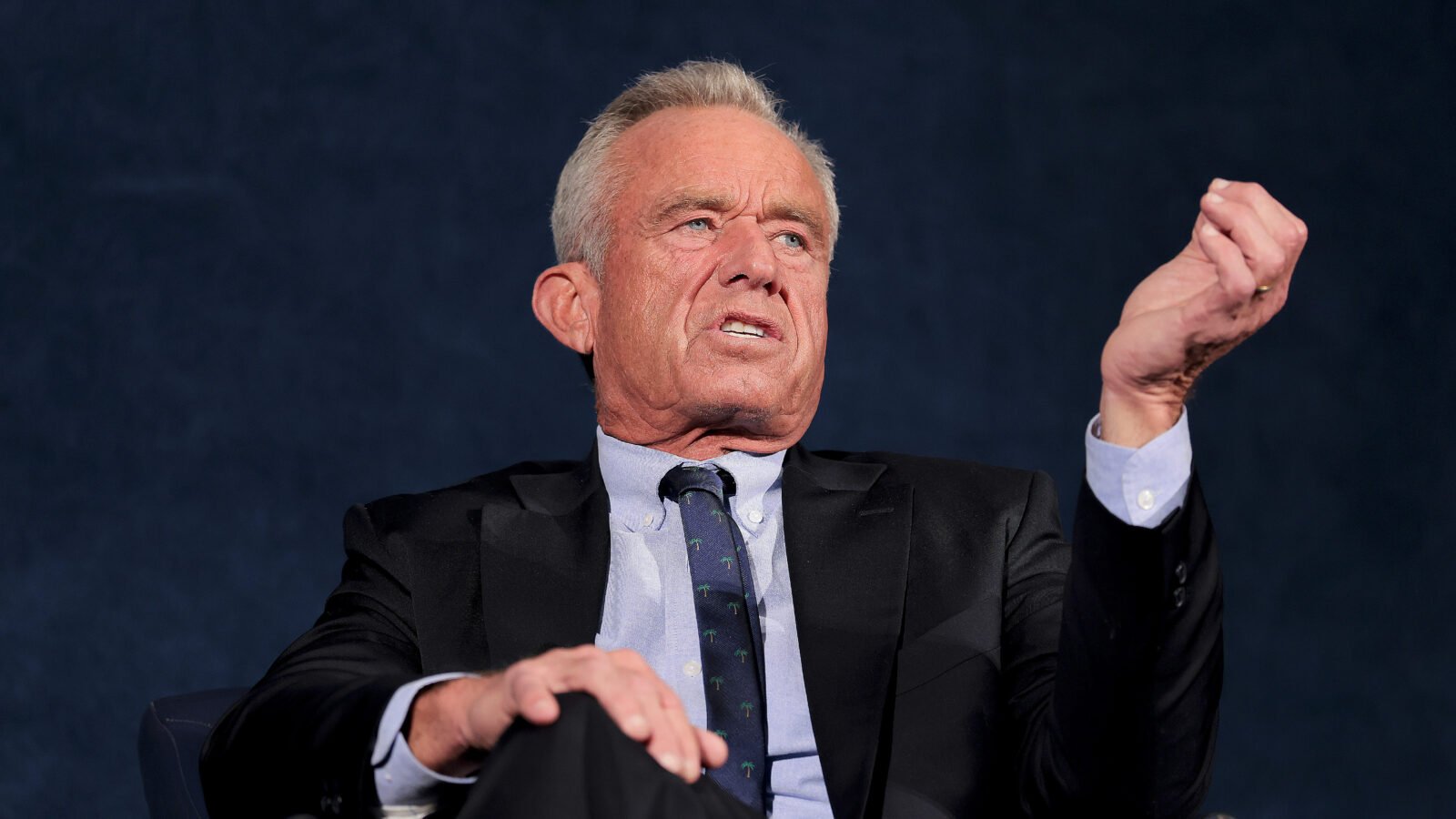 Kennedy sits in a suit speaking at Monday's announcement, holding his hand up.
