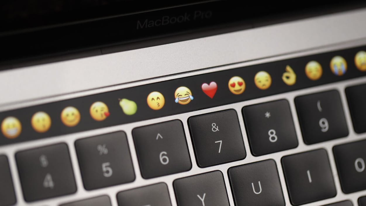 A close up of a keyboard with a row of emojis in the touch screen tool bar at the top.