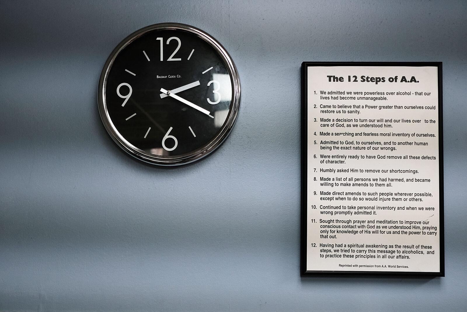 A clock hangs on the wall next to a framed list of the 12 steps of AA.