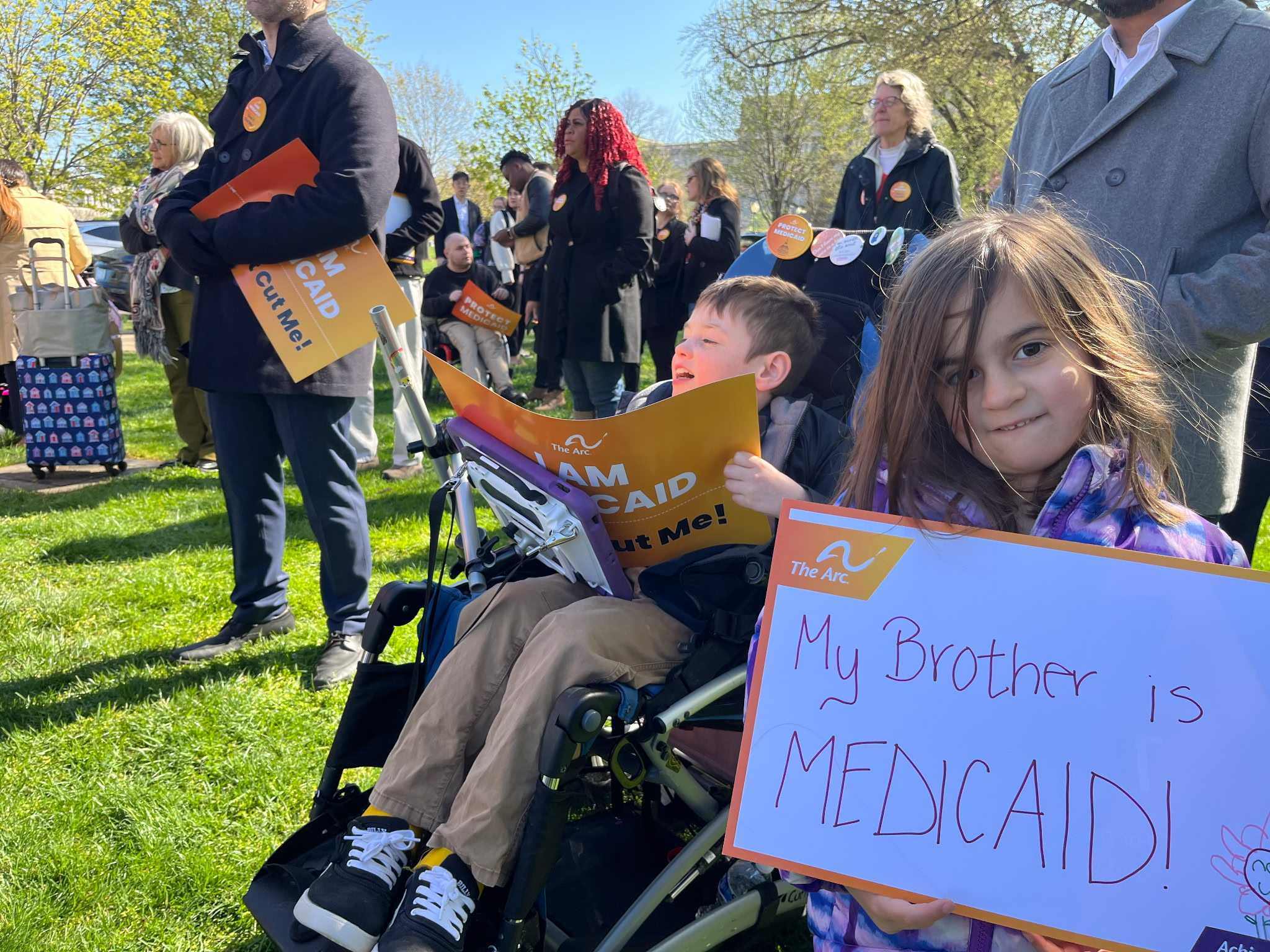 A young child with long hair looks right at the camera and holds up a sign that says "my brother is Medicaid!" She's next to a child that is presumably her brother, who has short hair and sits in a wheelchair and holds a sign saying "I am Medicaid."