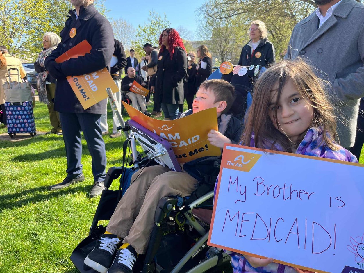 A young child with long hair looks right at the camera and holds up a sign that says "my brother is Medicaid!" She's next to a child that is presumably her brother, who has short hair and sits in a wheelchair and holds a sign saying "I am Medicaid."