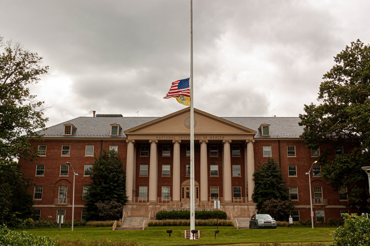 Building 1 of the National Institutes of Health in Bethesda, MD. It's a tall brick building, with a flagpole directly centered on the grass in front of it.