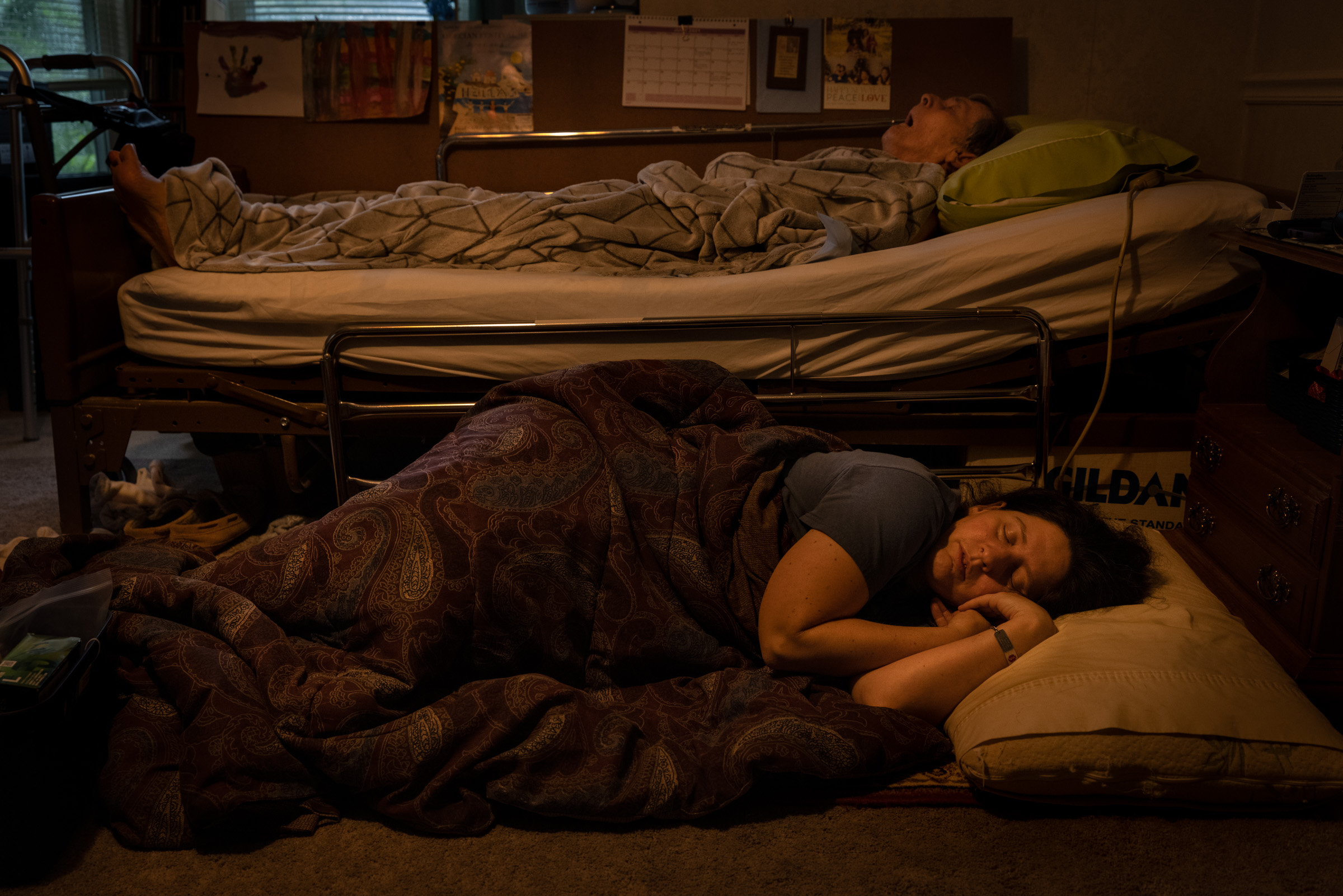 Leandra Manos sleeps on the floor below her father George in bed. Leandra is the caregiver for her father, who has dementia.