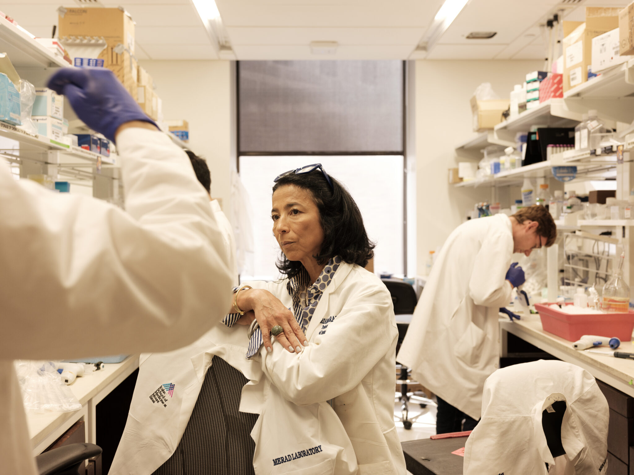 Miriam Merad stands stoically in a white coat in her lab. Multiple scientists work around her. Lots of motion but she is stable!
