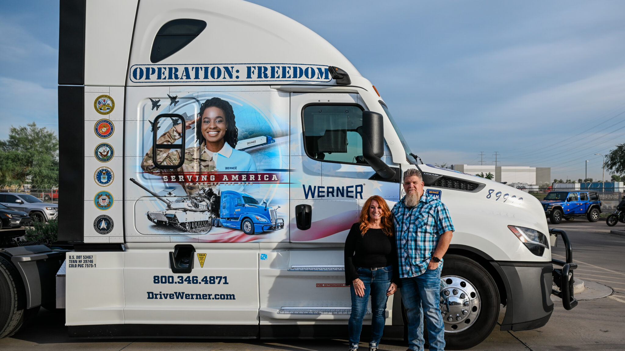 Gina and Steve stand in front of their truck. They both wear jeans, while Gina wears a longsleeve black blouse and Steve wears a blue shortsleeved plaid shirt. The truck says "Operation: Freedom" at the top and also "serving America" underneathe, against an illusustration of a saulting army person and a tank combined with a truck. 
