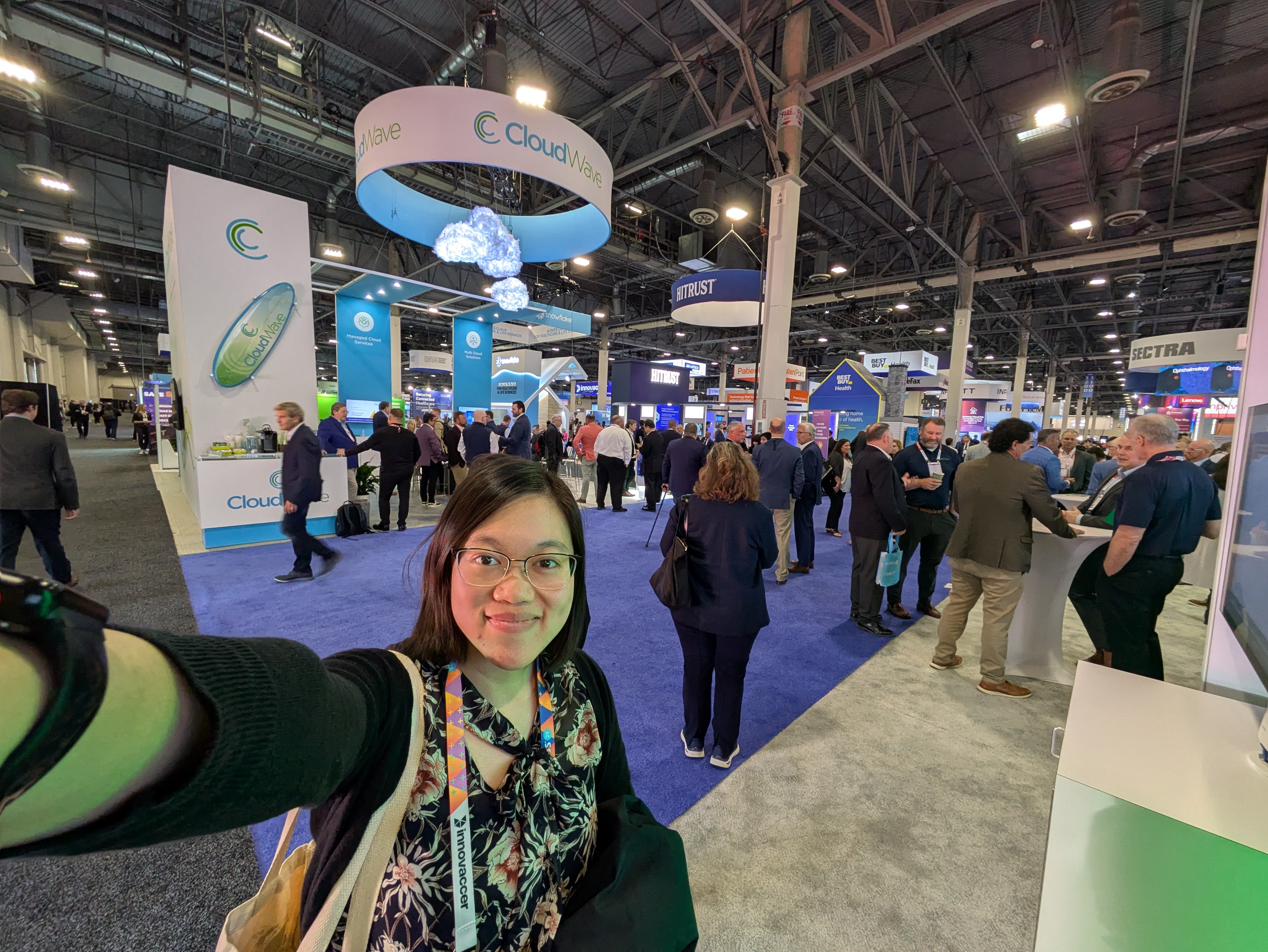 An Asian woman takes a selfie on the showroom floor of a conference, with many people behind her and companies' booths and signage visible in the background.