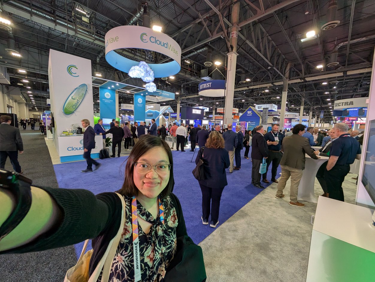 An Asian woman takes a selfie on the showroom floor of a conference, with many people behind her and companies' booths and signage visible in the background.