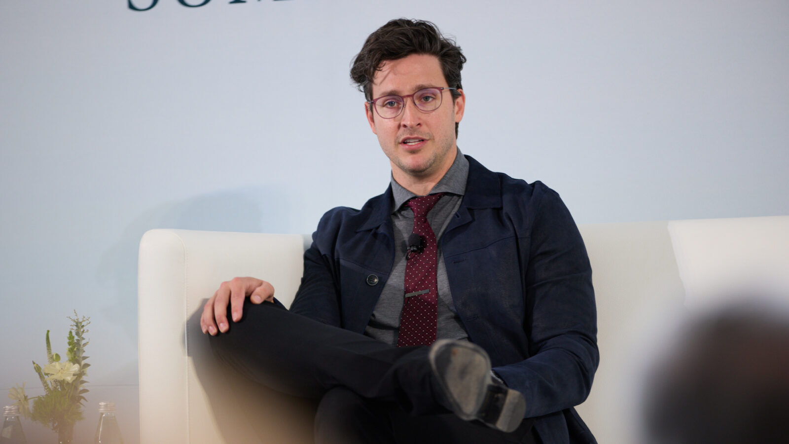 Doctor Mike sits in a white chair on stage at the STAT Summit in a blue grey suit with a grey shirt and red polka dot tie. One leg is propped up on the other. 