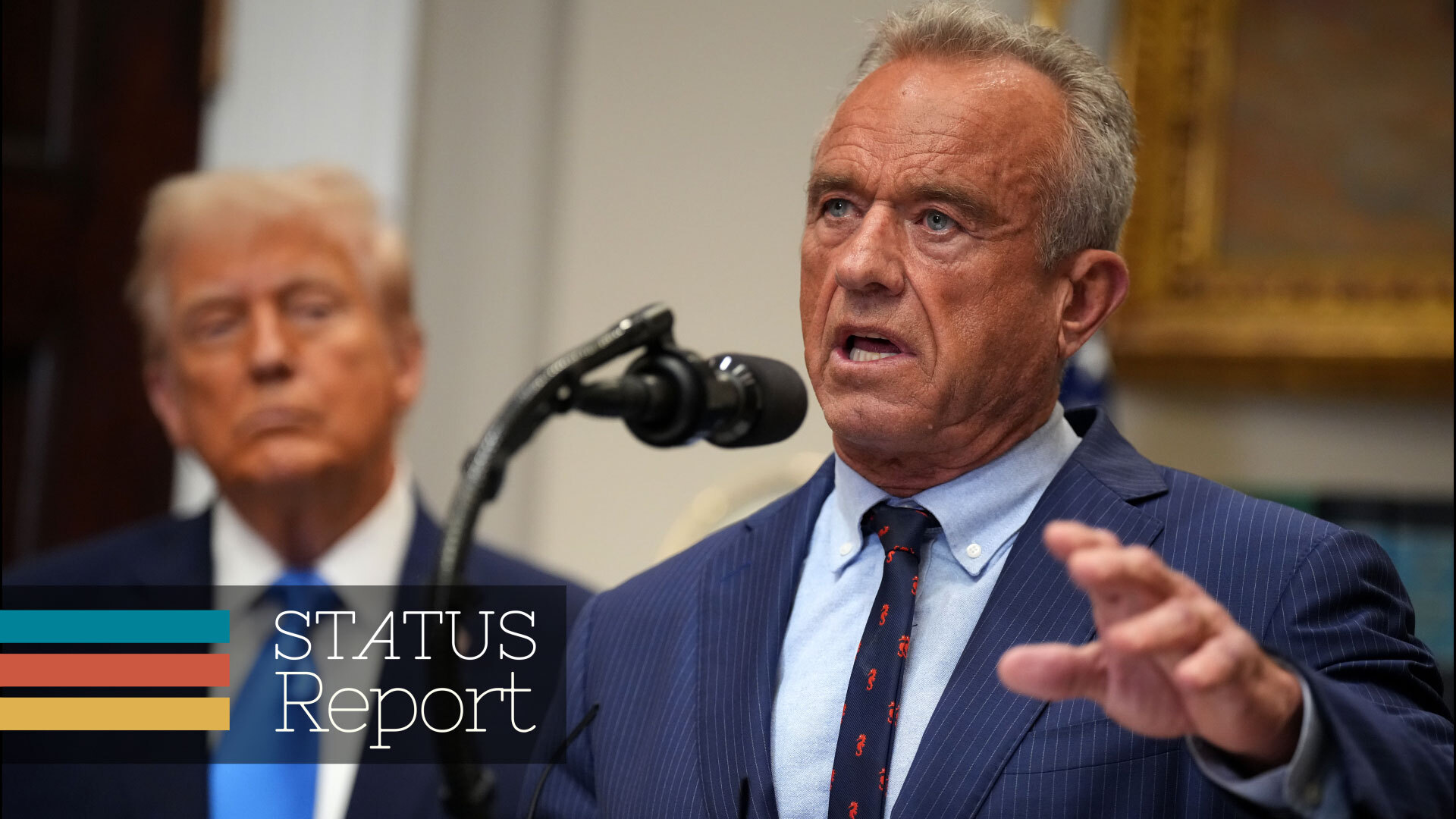 RFK Jr. stands at a microphone, speaking. President Trump is blurred in the background behind him. 