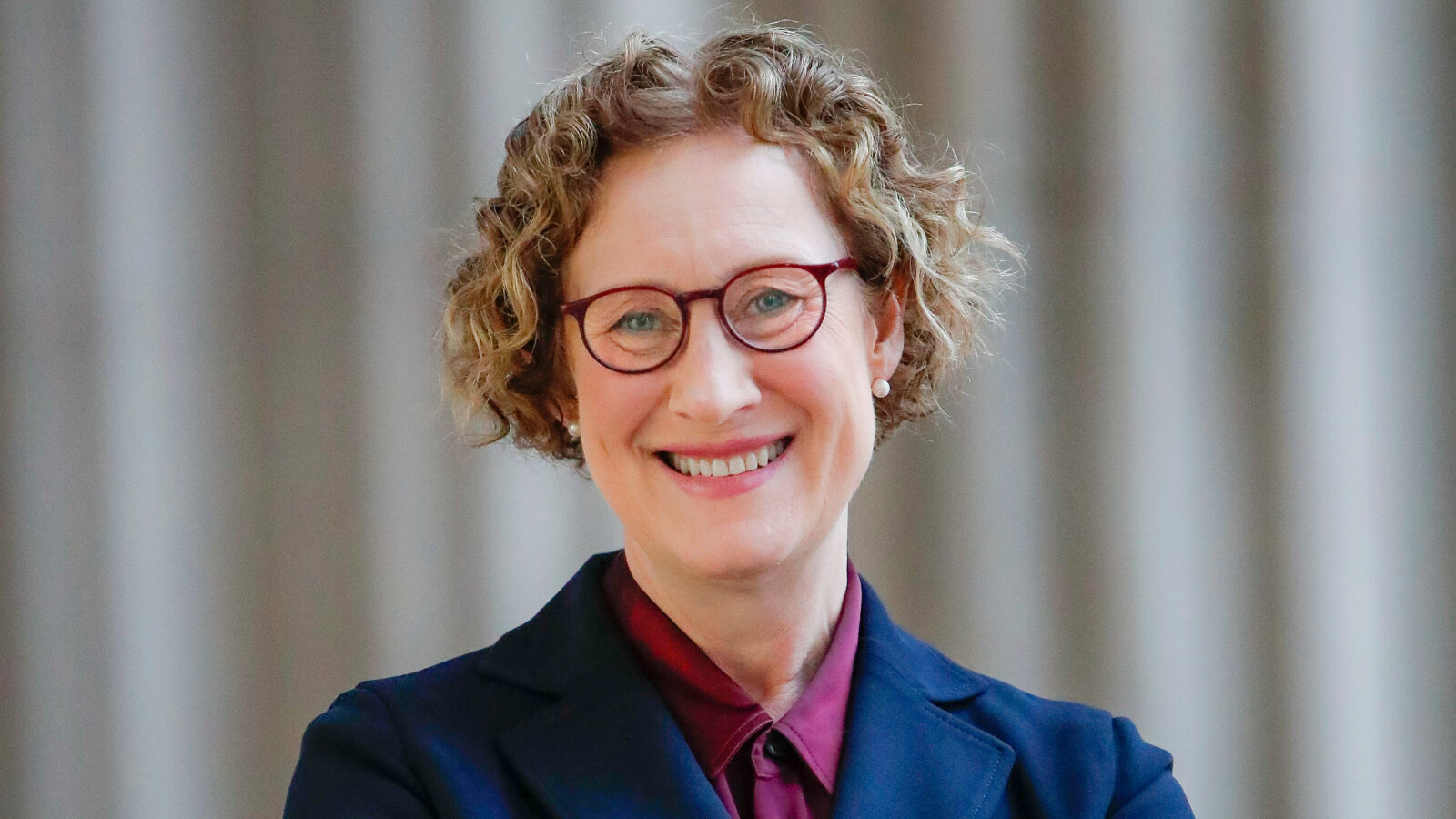 A portrait of Rebecca Cunningham, who has short, curly dirty blonde hair and wears round red glasses.