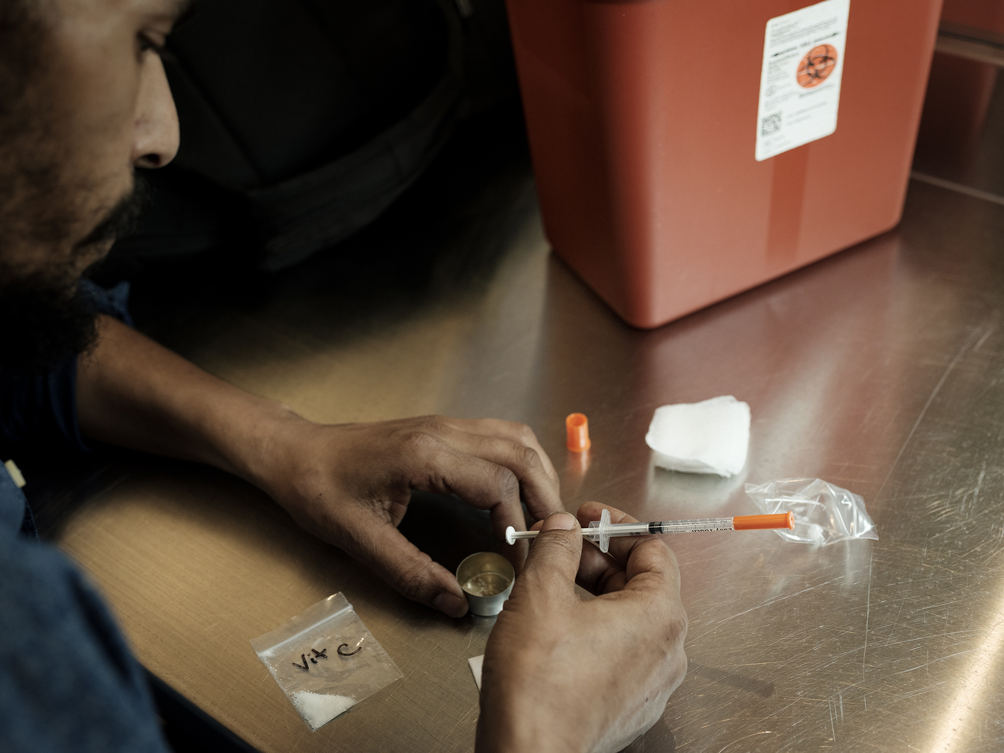 An over-the-shoulder close-up of a bearded man mixing cocaine and heroin in a small cup, with the back of a syringe, on a clean metal table.