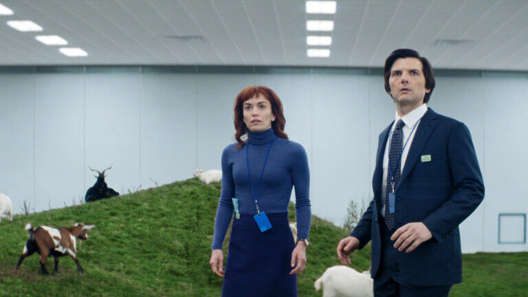 A screenshot from the tv show "Severance," in which two characters, a lady with amazing red hair microbangs and a plain guy in a suit, stand on guard in front of a bunch of grass and cows (?) that seem to be inside a warehouse. 