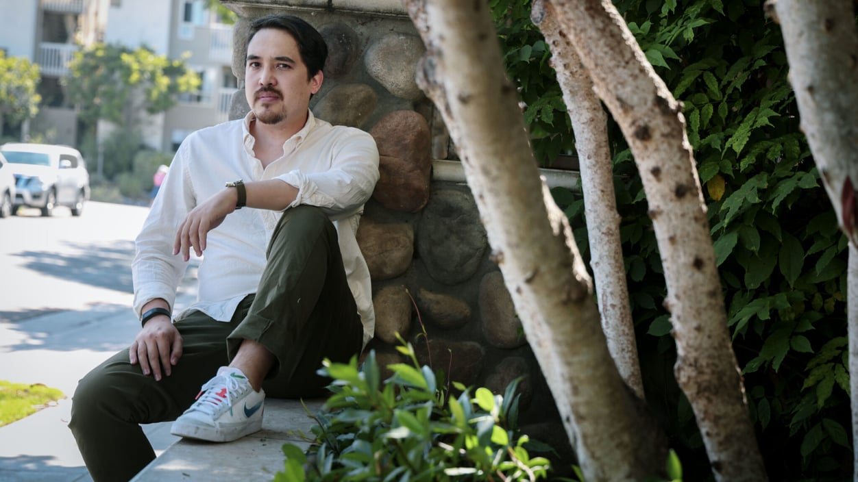 Vince Kato sits against a stone wall, next to a tree, in the shade.