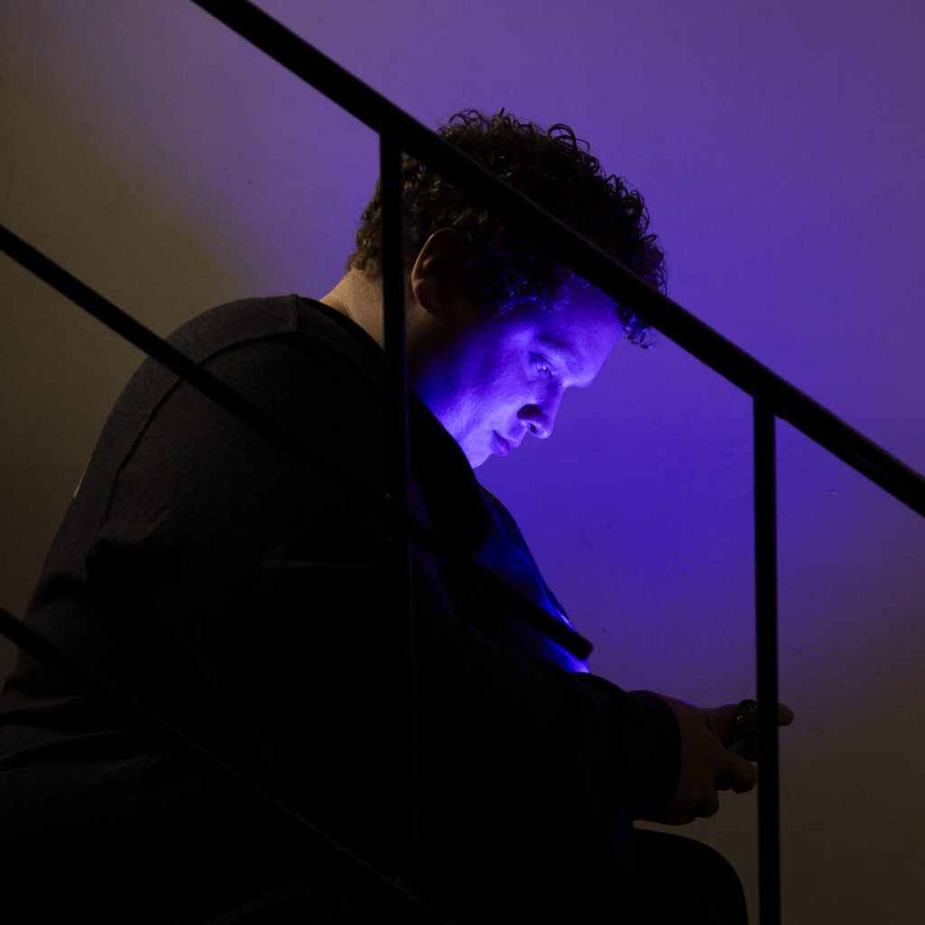A young man with curly hair sits on stairs, looking at a personal device, maybe a phone. He's illuminated by purple light. 